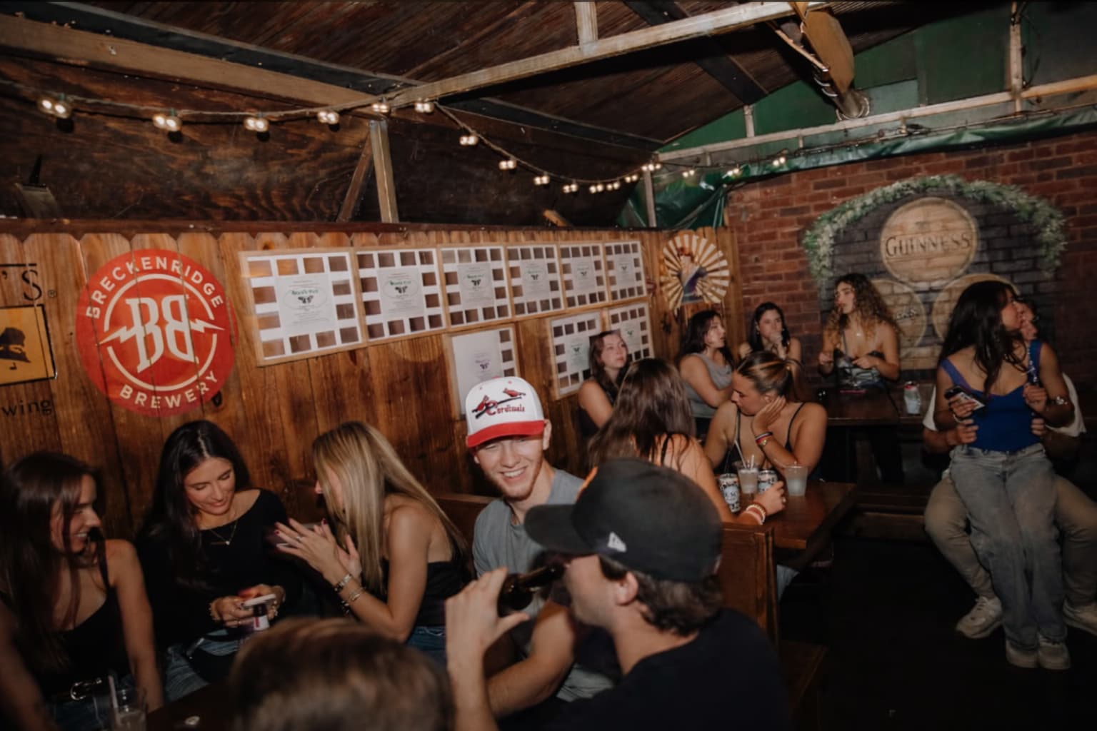 A packed patio crowd under string lights, Guinness mural on the brick wall