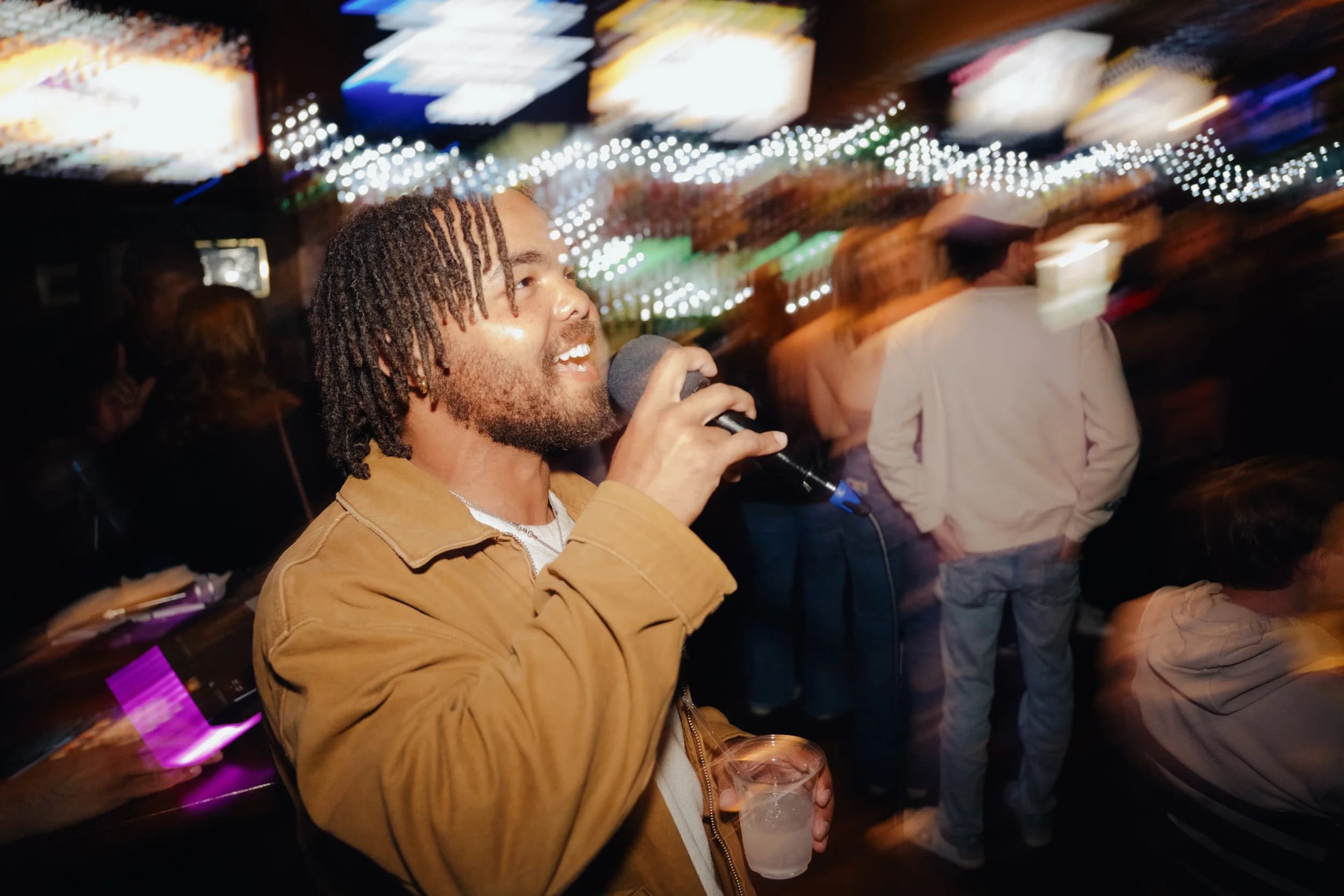 A singer mid-song at karaoke night, string lights and TVs glowing behind him